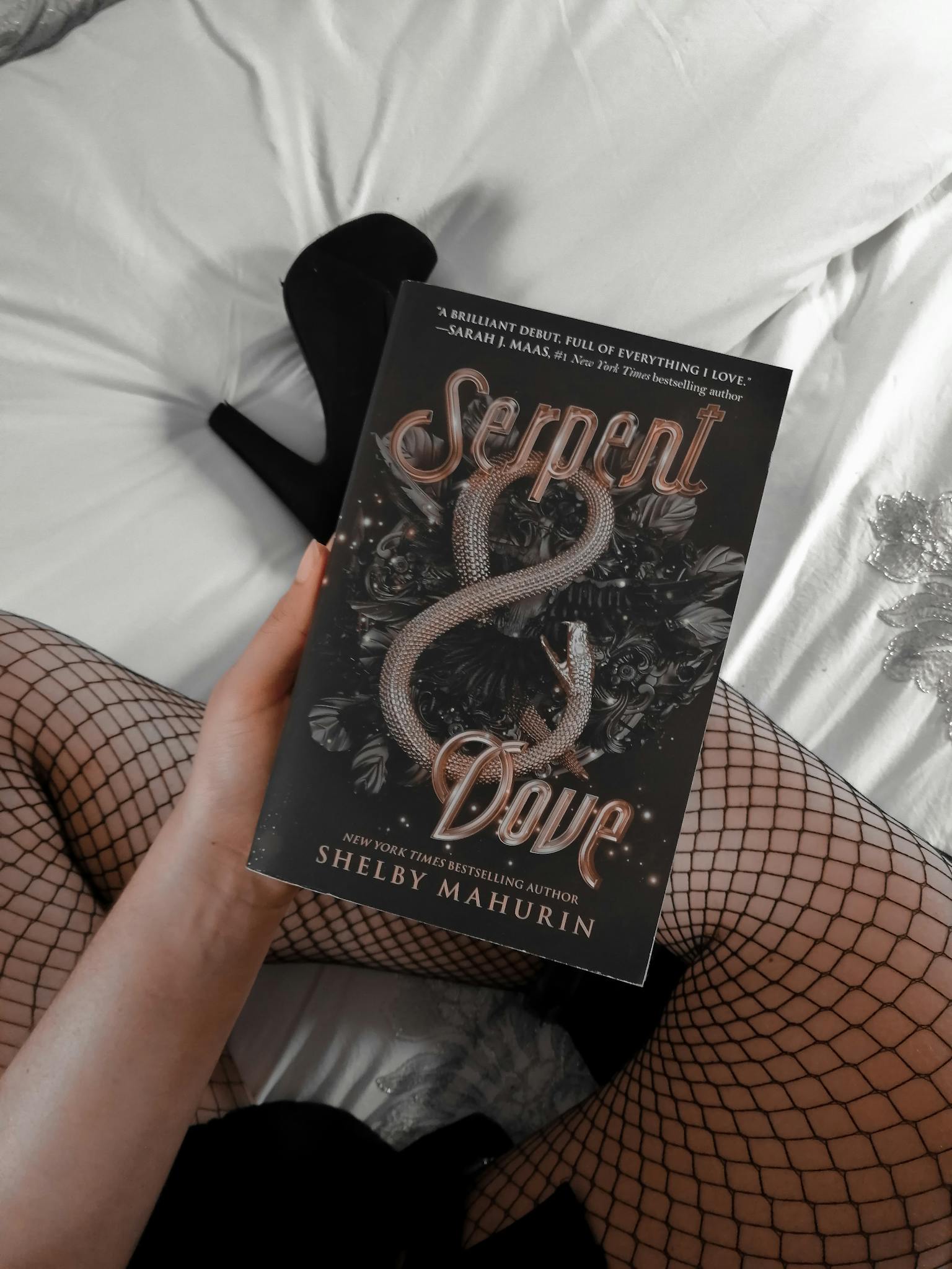 A person holds the novel 'Serpent & Dove' while wearing fishnet stockings, sitting on a bed.